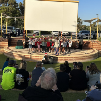 Children perform on an outdoor stage in front of a large screen, while a seated audience watches during a festive Christmas concert event.